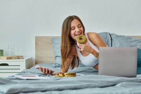 Photo for Joyous beautiful woman with long hair holding phone and enjoying donut while working from home - Royalty Free Image