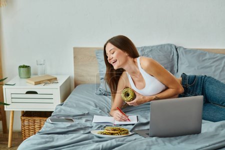 Photo for Cheerful attractive woman in homewear with long hair eating delicious donut while working from home - Royalty Free Image