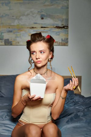 schöne junge Frau in beige sexy Dessous mit Korsett mit Lockenwicklern essen leckere Nudeln