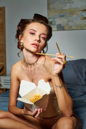 attraktive junge Frau mit beigem Korsett auf dem Bett sitzend mit einer Schachtel Nudeln und Essstäbchen in der Hand