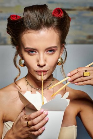 attractive young woman with hair curlers eating tasty noodles from box and looking at camera