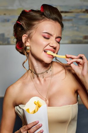 attraktive junge Frau mit Lockenwicklern in sexy beigem Korsett posiert emotional mit Pommes in den Händen