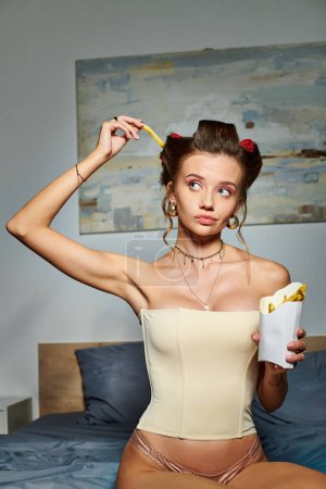 nachdenkliche Frau im sexy beigen Korsett mit Lockenwicklern, die mit Pommes in Bewegung posieren und wegschauen