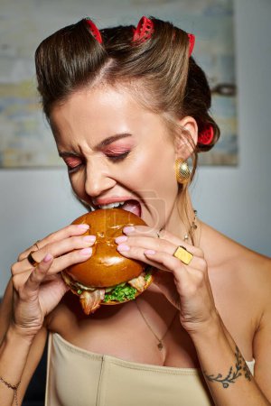 joven hermosa mujer con rizadores de pelo y accesorios comiendo deliciosa hamburguesa con apetito