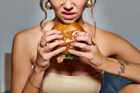 Téléchargez les photos : Vue recadrée de jeune femme émotionnelle avec accessoires et tatouage à portée de main manger énorme hamburger - en image libre de droit