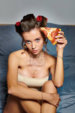attraktive Frau mit Lockenwicklern und Accessoires posiert mit einem Stück Pizza und blickt in die Kamera