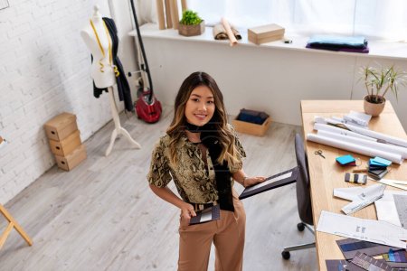 fröhliche asiatische Designerin mit Farbmustern vor der Kamera in modernem Atelier, Hochwinkelaufnahme