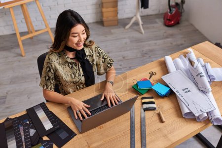 vue grand angle de joyeux asiatique styliste regardant ordinateur portable près des échantillons de couleur sur le bureau en atelier