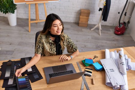 glückliche asiatische Modedesignerin lächelt neben Laptop und Farbmustern im Atelier