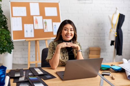 fröhliche asiatische Stylistin blickt in die Kamera in der Nähe von Laptop und Farbmustern auf dem Schreibtisch im Atelier