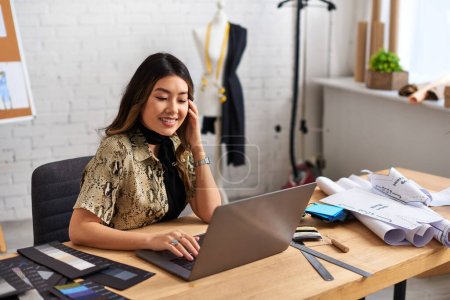 glückliche asiatische Modedesignerin arbeitet im eigenen Atelier an Laptop in der Nähe von Nählatten und Uhren