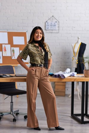 fröhliche und trendige asiatische Designerin posiert mit den Händen in den Taschen am Schreibtisch im privaten Atelier