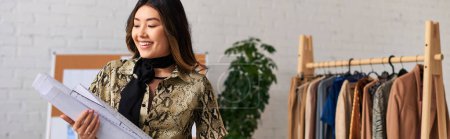 Joyeux concepteur de vêtements asiatiques tenant des motifs de couture dans son propre atelier, entreprise de mode, bannière