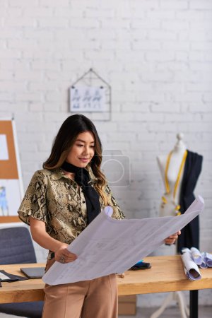 diseñador asiático positivo mirando patrones de costura cerca de escritorio de trabajo en estudio de moda privada