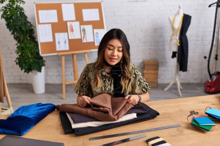 Téléchargez les photos : Heureux asiatique personnel styliste contrôle qualité de tissus échantillons sur bureau dans propre atelier - en image libre de droit