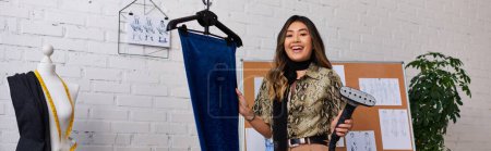 young asian designer with steamer and velour fabric smiling at camera in personal atelier, banner
