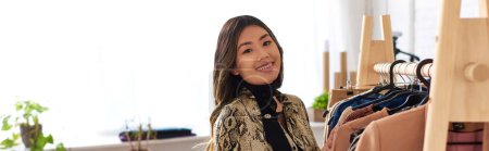 Téléchargez les photos : Heureux asiatique personnel styliste regarder caméra près de rack avec des vêtements à la mode dans atelier, bannière - en image libre de droit