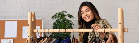 Téléchargez les photos : Joyeuse couturière asiatique souriant à la caméra près de rack avec des vêtements sur mesure dans son propre atelier, bannière - en image libre de droit