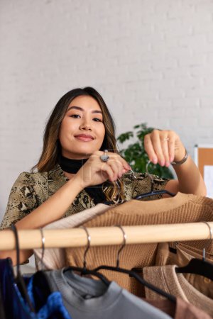 Téléchargez les photos : Attrayant heureux asiatique styliste avec à la mode sur mesure vêtements regardant caméra près rack dans propre studio - en image libre de droit