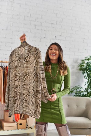 overjoyed asian stylist holding trendy animal print shirt and smiling at camera in fashion studio