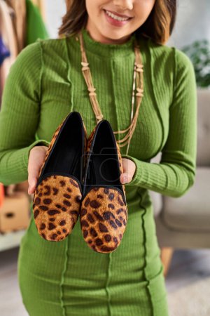 Téléchargez les photos : Vue recadrée d'une femme souriante à la mode montrant des chaussures d'impression animale à la mode dans un atelier de vêtements - en image libre de droit