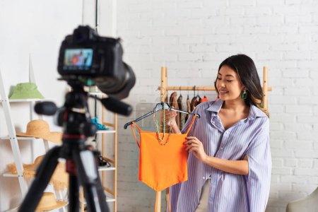freudige asiatische Modedesignerin zeigt Crop Top und Accessoires während Videoblog nahe Digitalkamera