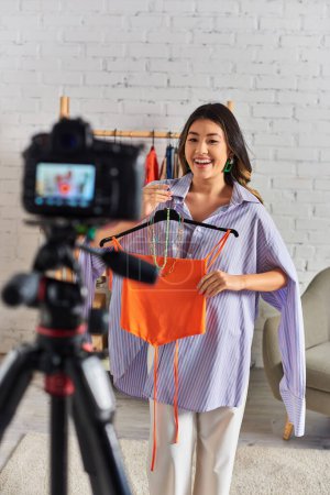 Téléchargez les photos : Joyeux asiatique styliste tenant crop top et accessoires près appareil photo numérique pendant mode vlog - en image libre de droit
