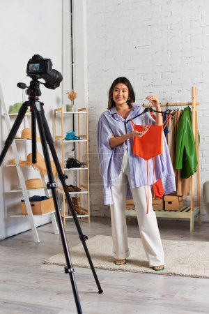 Téléchargez les photos : Mode asiatique styliste tenant crop top et accessoires en face de l'appareil photo numérique pendant blog vidéo - en image libre de droit