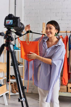 Téléchargez les photos : Souriant asiatique styliste montrant avec crop top et accessoires devant appareil photo numérique dans son propre studio - en image libre de droit