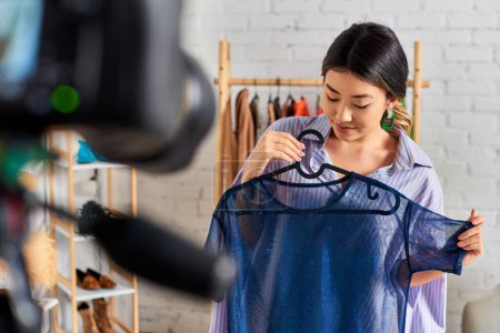 Téléchargez les photos : Jeune couturière asiatique avec chemisier transparent à la mode près de l'appareil photo numérique flou en atelier - en image libre de droit