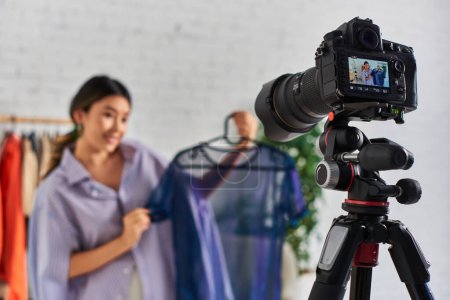 Téléchargez les photos : Focus sur appareil photo numérique près de jeune styliste asiatique avec chemisier tendance en atelier, mode vlog - en image libre de droit