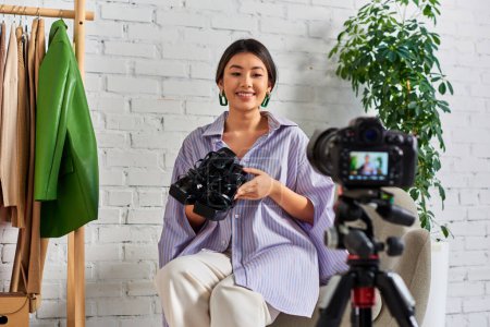 Téléchargez les photos : Sourire asiatique couturière avec des chaussures à la mode en face de l'appareil photo numérique pendant blog vidéo de mode - en image libre de droit