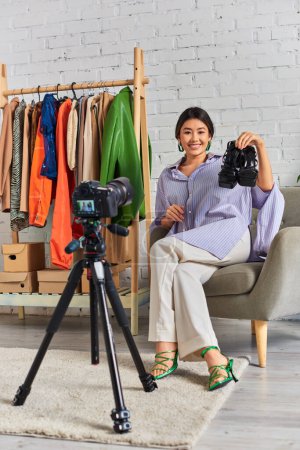 Téléchargez les photos : Positif asiatique styliste de mode montrant sandales à la mode pendant blog vidéo dans l'atelier de vêtements personnels - en image libre de droit