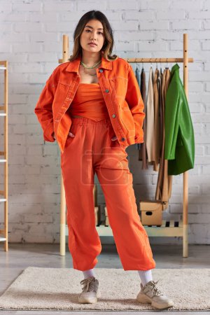 junge asiatische Modedesignerin in leuchtend orangefarbener Kleidung posiert mit Händen in Taschen im eigenen Atelier