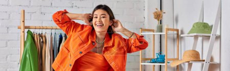 encantado diseñador de moda asiático posando en ropa naranja brillante en el estudio, bandera horizontal