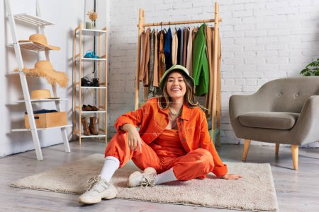 Téléchargez les photos : Souriant asiatique styliste dans lumineux vêtements et panama chapeau assis sur sol dans personnel atelier de mode - en image libre de droit