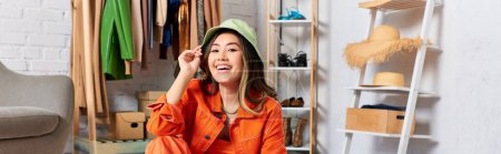 fröhliche asiatische Stylistin posiert in orangefarbener Kleidung und Panamahut im Atelier, Modegeschäft, Banner