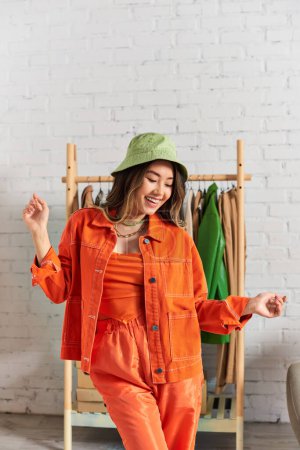 glückliche asiatische Stylistin posiert in orangefarbener Kleidung und Panamahut in privatem Atelier, Modegeschäft