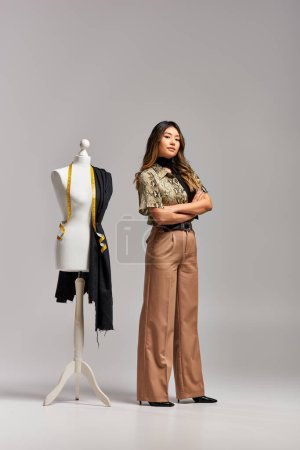 Téléchargez les photos : Designer asiatique créatif avec bras croisés près du mannequin avec tissu et ruban à mesurer sur gris - en image libre de droit