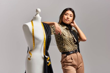 créatif asiatique designer regarder loin près mannequin avec tissu et ruban à mesurer sur gris