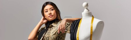 styliste asiatique moderne regardant loin près mannequin avec tissu et ruban à mesurer sur gris, bannière