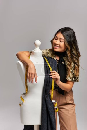 zufriedene asiatische Designerin posiert in der Nähe einer Schaufensterpuppe mit Stoff und Maßband auf grauem Hintergrund