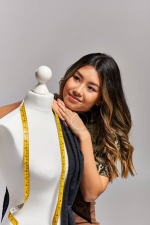rêveuse asiatique vêtements designer regarder loin près de mannequin avec ruban à mesurer sur fond gris