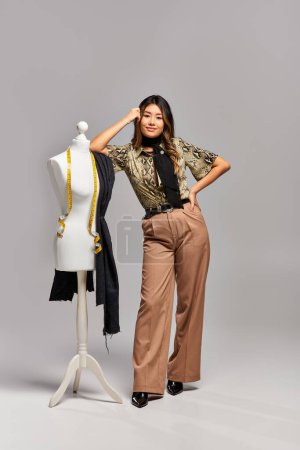 trendy asian fashion designer posing near mannequin with measuring tape and fabric on grey backdrop