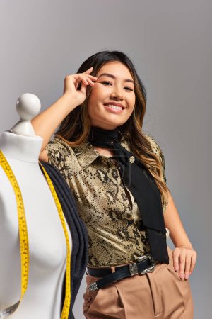 sourire asiatique créateur de mode avec ruban à mesurer près de mannequin avec tissu sur fond gris