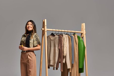 heureux asiatique styliste avec mesure ruban regarder loin près de rack avec sur mesure vêtements sur gris