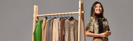 ravi designer asiatique regardant loin près de rack avec des vêtements à la mode sur fond gris, bannière