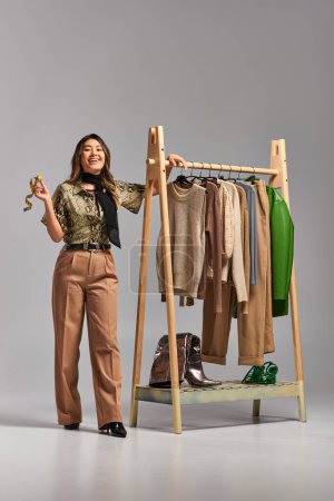 Photo for Cheerful asian stylist with measuring tape looking at camera near rack with bespoke clothes on grey - Royalty Free Image