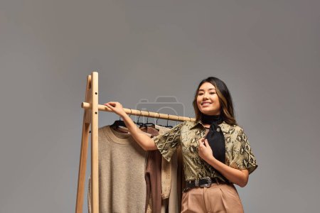 Photo for Young happy asian clothes stylist posing near rack with trendy bespoke clothes on grey backdrop - Royalty Free Image
