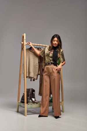 modische asiatische Kleidungsdesignerin steht in der Nähe des Regals mit trendigen maßgeschneiderten Kleidern vor grauem Hintergrund